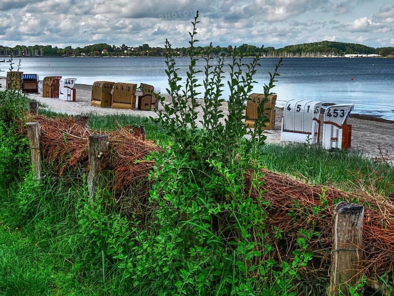 HDR image of beach chairs on the beach, grasses and wood in the foreground, cloudy sky in the background, beach chairs on a quiet sandy beach by the sea, clouds over the sea, Eckernfoerde, germany