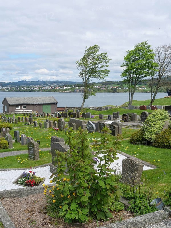 Cemetery with many gravestones, in the background a body of water, trees and a small house under a cloudy sky, old stone church and many gravestones by the water with ships in the background, haugesund, norway