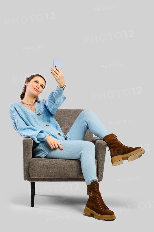 A passionate woman in a blue knitted outfit is seated comfortably on a chair, engaging with her smartphone. The image captures a moment of leisure and connection