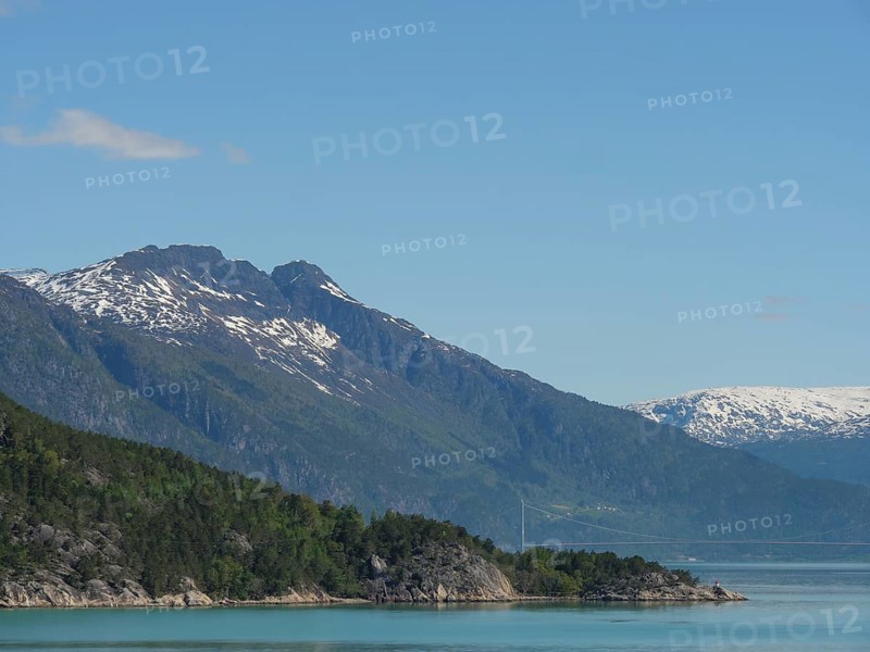 View of steep, forested mountains, a lake and snow-capped peaks in sunny weather, greenish shimmering water in a fjord with snow-capped mountains and green trees, eidfjord, Norway, Europe