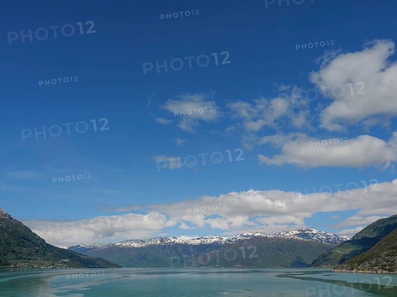 Expansive view of a mountain range under a clear blue sky with some clouds and a calm lake, greenish shimmering water in a fjord with snow-capped mountains and green trees, eidfjord, Norway, Europe