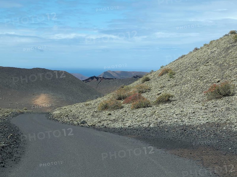A tarmac road winds its way through overgrown, barren hills under a slightly cloudy sky, barren landscape with roads, craters and small bushes, Lanzarote, spain