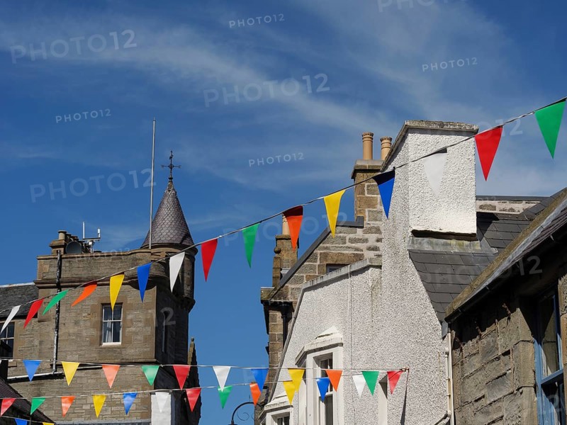 Street scene with colourful pennants hanging between buildings and a tower, calm sea with rocks and fields with historic houses, Lerwick, Shetlands, Scotland, United Kingdom, Europe