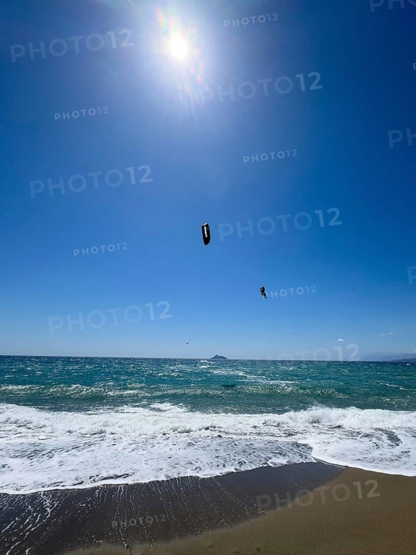 Kitesurfer Kitesurfer takes off in strong wind and waves rough sea moving sea flies with inflated sail high through the air towards the sun, in front beach with white small whitecaps, Kalamaki, Crete, Greece, Europe