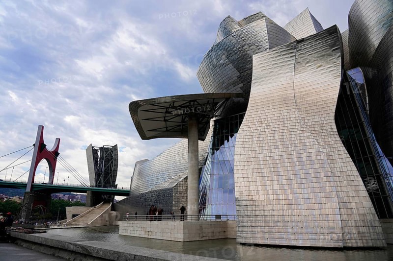 Guggenheim Museum Bilbao, Spain, Europe, The picture shows modern metallic buildings next to a red bridge under a cloudy sky, Guggenheim Museum Bilbao on the banks of the river Nervion, architect Frank O. Gehry, Bilbao, Basque Country, Europe