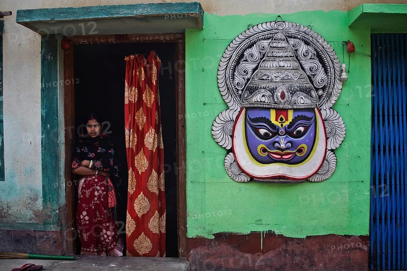 Tall mask representing a Kathakali dancer hanging on the outer wall of the house of a craftsman, craftsman making and selling Purulia Chhau dance masks, woman standing at the entrance of her house, West Bengal, India, Asia