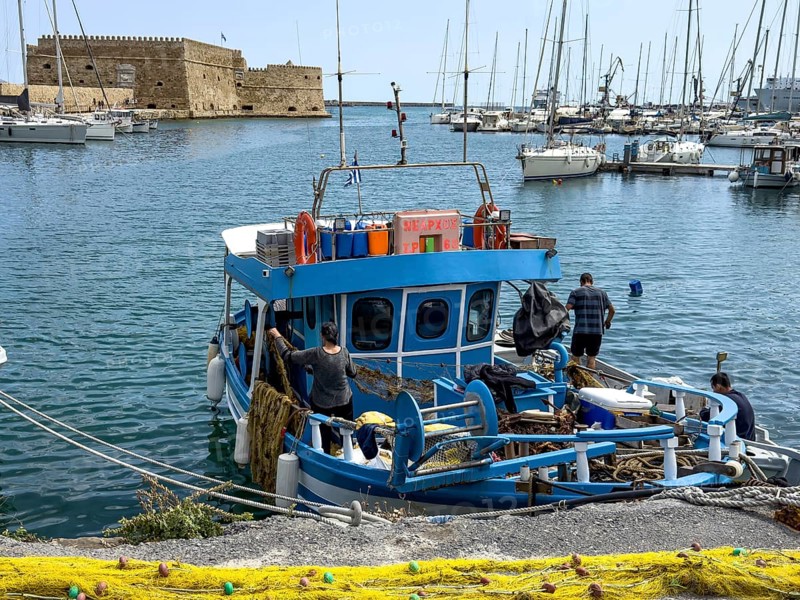 In the foreground yellow fishing net from behind local Greek fishermen arranging their nets on local small fishing boat, in the background historical fortress built in Middle Ages 16th century by Republic of Venice old name Castello Rocca al Mare today Koules in harbour of Iraklion, Heraklion, Crete, Greece, Europe