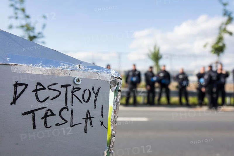 Destroy Tesla! slogan on a cardboard Cybertruck model in front of the factory premises guarded by police officers at the Water. Forest. Justice against the expansion of the Tesla Gigafactory in Gruenheide near Berlin, 11 May 2024