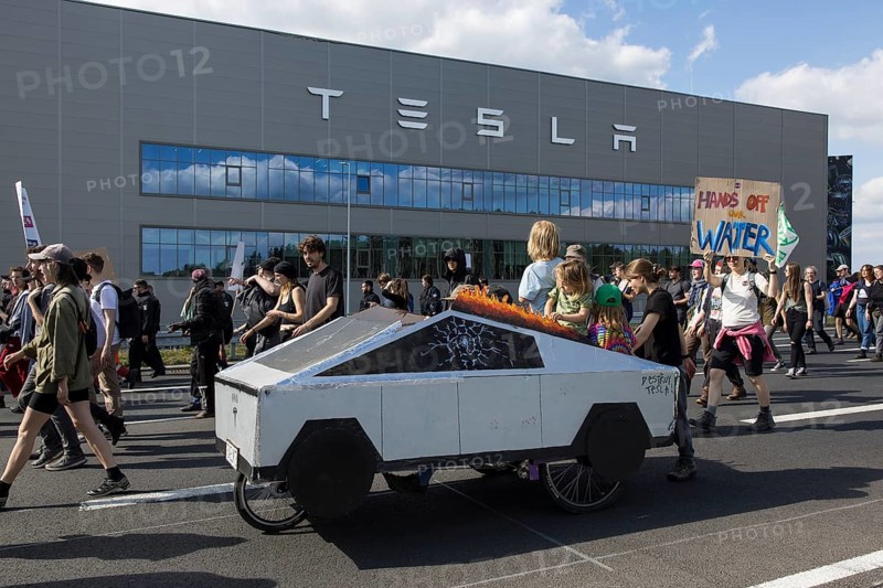 Participants with a Cybertruck cardboard model on a bicycle and sign Hands off our water in front of the Tesla Gigafactory at the demonstration Water. Forest. Justice against the expansion of the Tesla Gigafactory in Gruenheide near Berlin, 11 May 2024