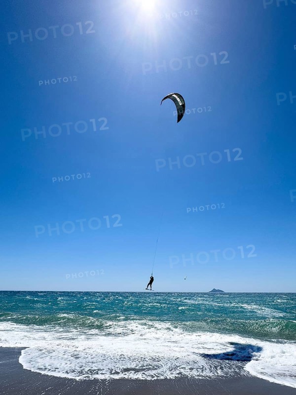 Kitesurfer Kitesurfer takes off in strong wind and waves rough sea moving sea flies with inflated sail high through the air towards the sun, in front beach with white small whitecaps, Kalamaki, Crete, Greece, Europe