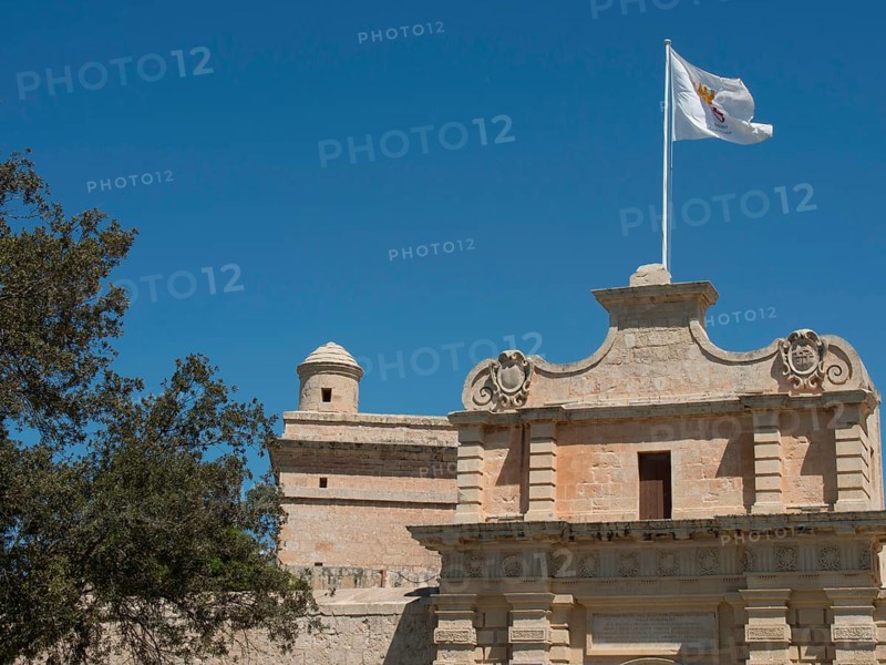 An old castle with a flag and a tower in the foreground, bright blue sky, historic buildings with beautiful windows, balconies and small towers, Mdina, Malta, Europe