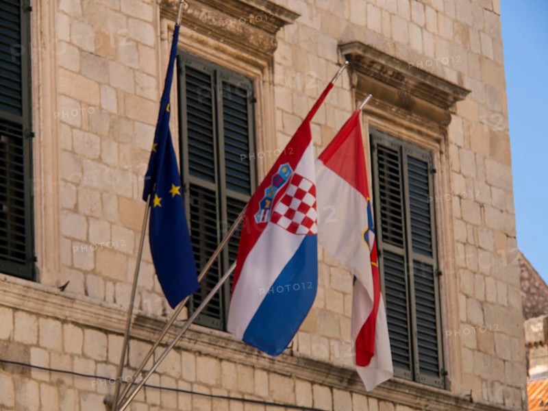 Three flags, including the European and Croatian flags, fly in front of a historic building with closed shutters, the old town of Dubrovnik with historic houses, churches, red roofs and fortress walls, dubrovnik, coratia