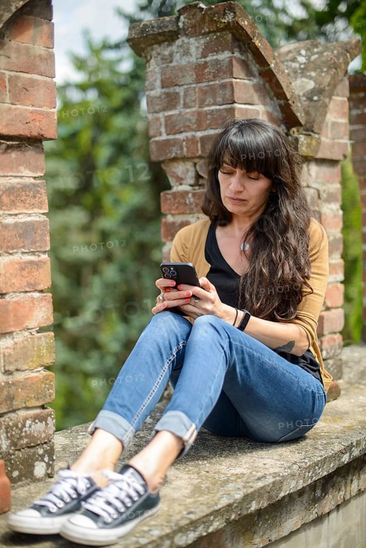 Young woman in casual clothing focused on conversation with smartphone in a peaceful garden with blurred brickwork in the foreground
