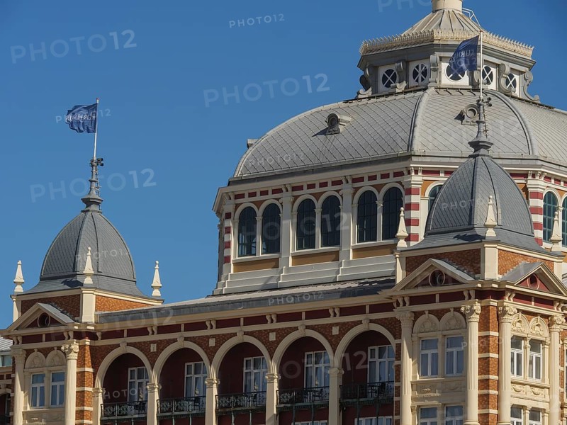 Historic building with domes and flags under a blue sky, parasols and a pier on the beach of Scheveningen, Scheveningen, Netherlands