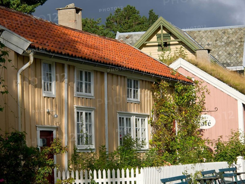Yellow and pink house with a cafe sign and lush vegetation, colourful wooden houses behind white wooden fences, Molde, Norway, Europe