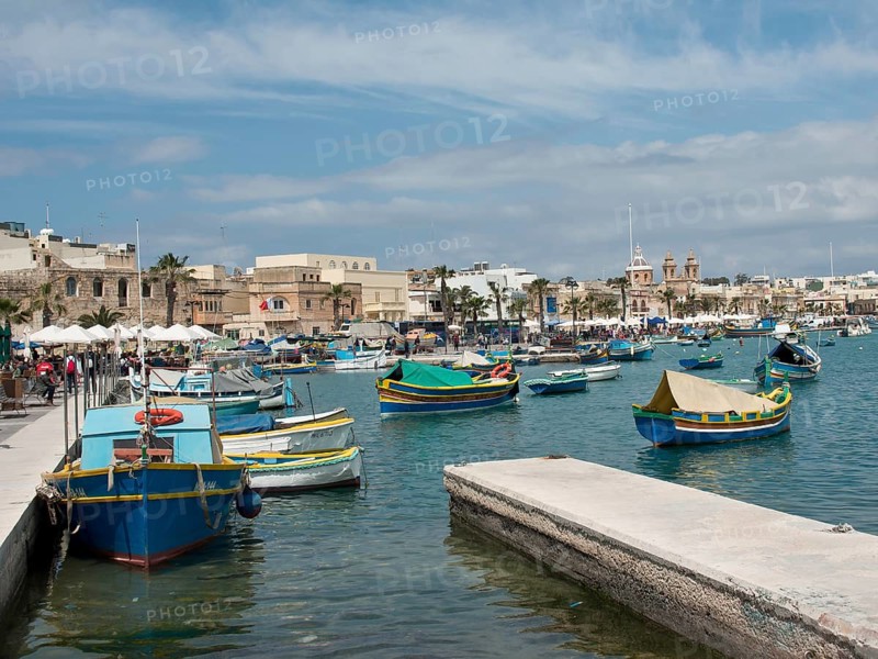 Several colourful boats in the harbour, surrounded by historic buildings and numerous palm trees under a blue sky, many colourful fishing boats in a small harbour against a historic backdrop, Marsaxlokk, malta