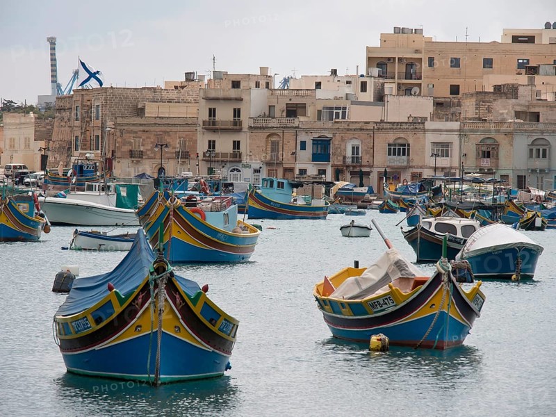 Several colourful boats in the harbour, behind them a town with many residential buildings and calm water, colourful boats in a historic harbour on the Mediterranean, Marsaxlokk, malta