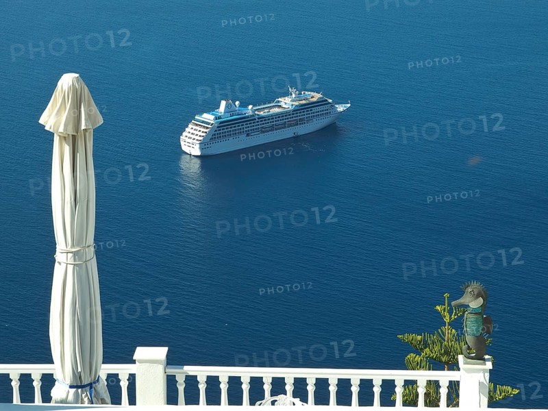 A cruise ship in the blue sea near a coastline with a parasol and railing in the foreground, The volcanic island of Santorini with blue and white houses and churches, flowers and the deep blue sea, Santorini, Greece, Europe