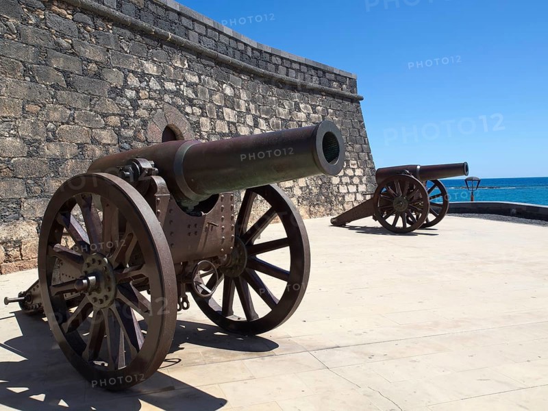 Two ancient cannons next to a fortress with a view of the sea, the island of Lanzarote with volcanic mountains, palm trees and wise houses, lanzarote, spain