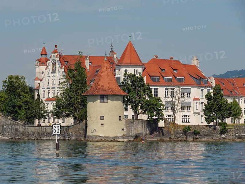 A castle with a red roof on the shore of a lake, a tower and dense trees, sky in the background, view of a lakeside town with lighthouse and church towers, Lindau, Germany, Europe
