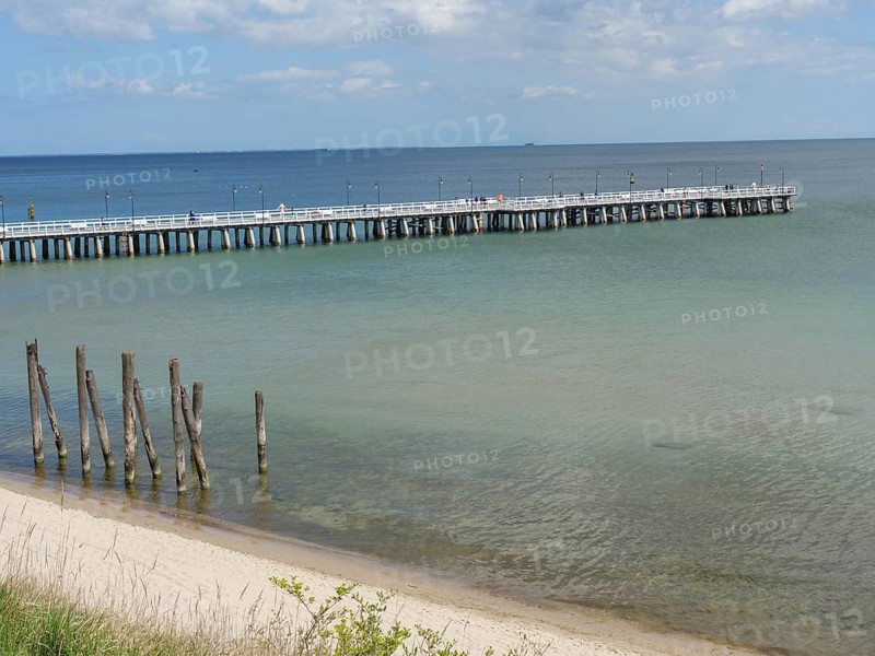 A long jetty leads into the calm sea under a cloudy sky on a sandy beach, spring on the Baltic coast with green fishing boats on the beach and green trees on the shore, Sopot, Poland, Europe