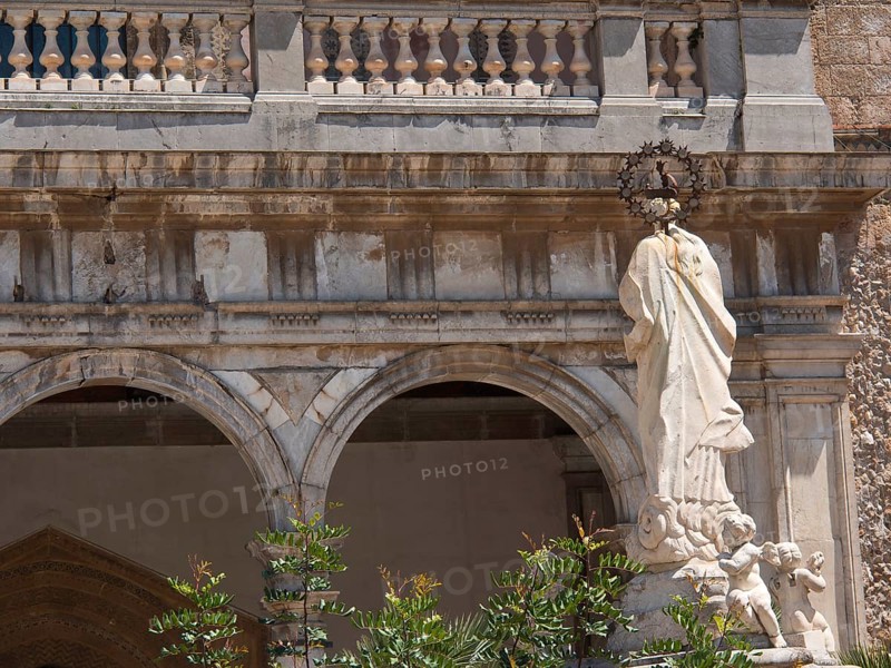 Marble sculpture of a figure and two angels in front of a church with arches and ornate details, palermo in sicily with an impressive cathedral, monuments and old houses with balconies, palm trees and flowers in italy, palermo, sicily