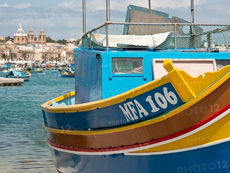 A blue and yellow fishing boat at a harbour with a coastal town in the background, many colourful fishing boats in a small harbour against a historical backdrop, Marsaxlokk, malta