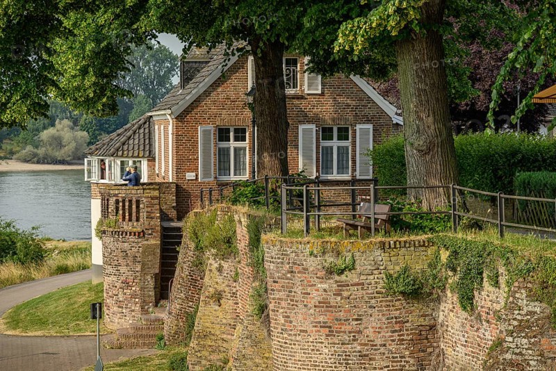 An old house on a riverbank, surrounded by a wall and trees, summery historical landscape, the promenade of Rees on the Rhine with green trees in spring, Rees, germany