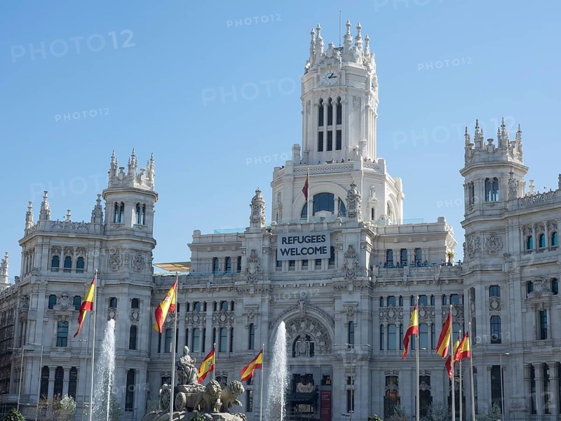 Monumental white palace with Spanish flags, spring fountain and a welcome banner for refugees, Madrid, Spain, Europe