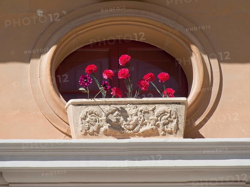 An oval window with a decorative flower box full of red flowers in front of it, the town of mdina on the island of malta with historic houses, colourful balconies, magnificent churches, mdina, Malta, Europe
