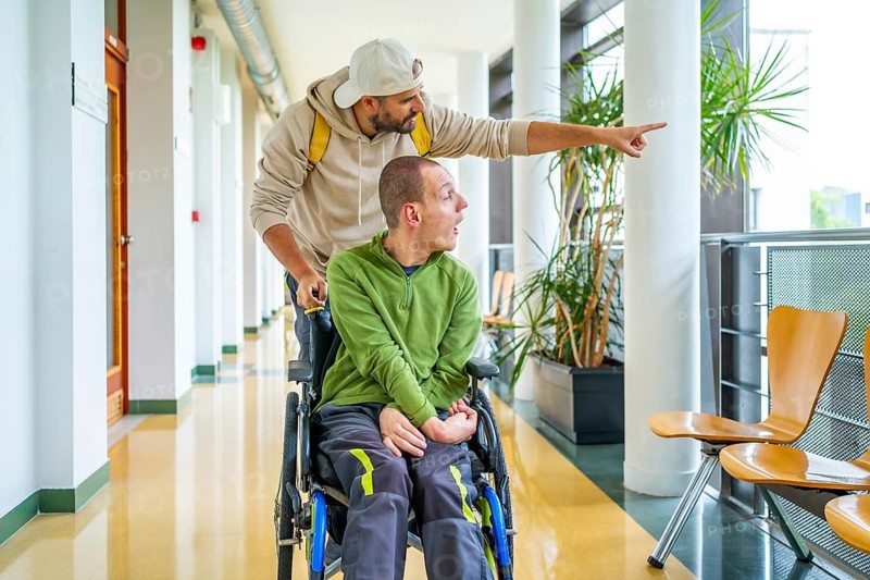 Caucasian young man showing something outside the university to a disabled friend sitting on wheelchair