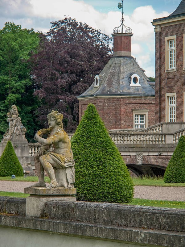 Stone sculpture in front of a historic castle with brick walls, green bushes and trees under a cloudy sky, historic castle with a green park with sculptures and paths, nordkirchen, germany