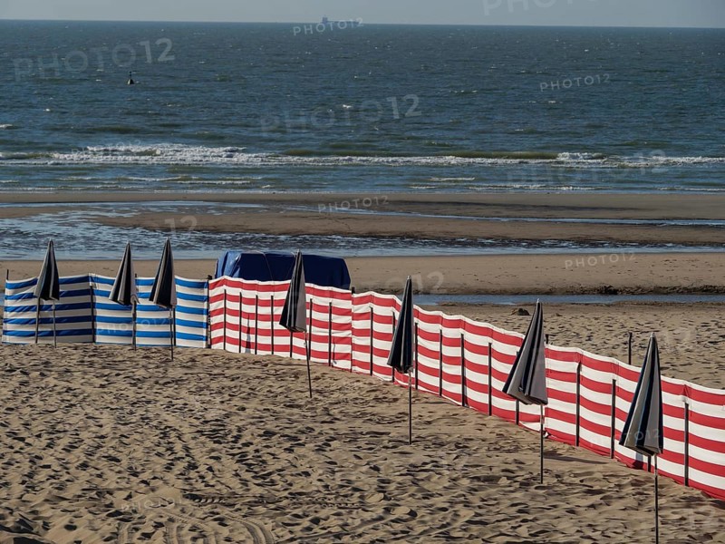 Wide sandy beach area with scattered blue and white striped and black umbrellas and the sea with waves in the background, colourful beach chairs by the sea with blue sky, de haan, belgium