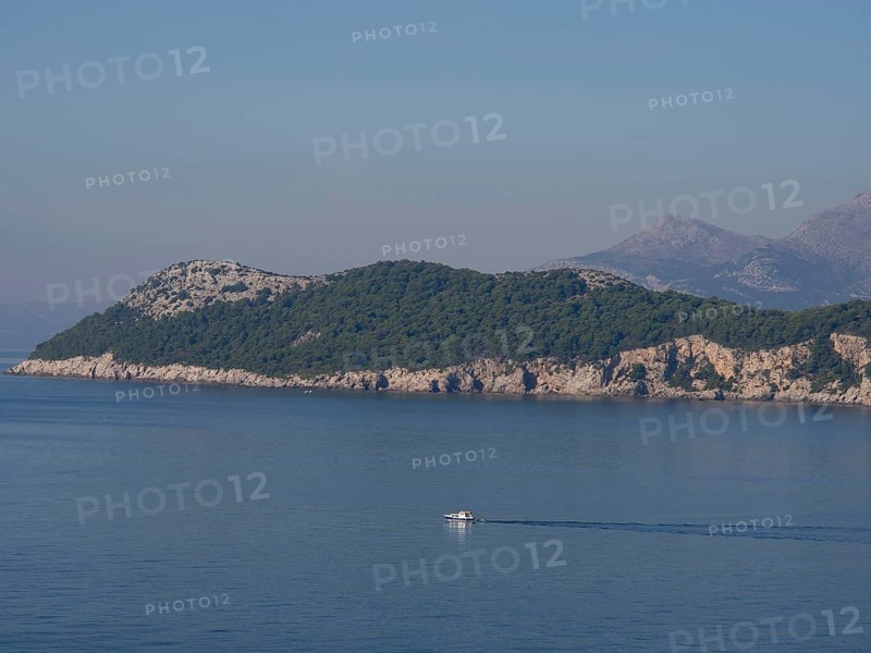 A small boat sails on the calm sea along a forested chain of islands, the old town of Dubrovnik with historic houses, churches, red roofs and fortress walls, dubrovnik, coratia