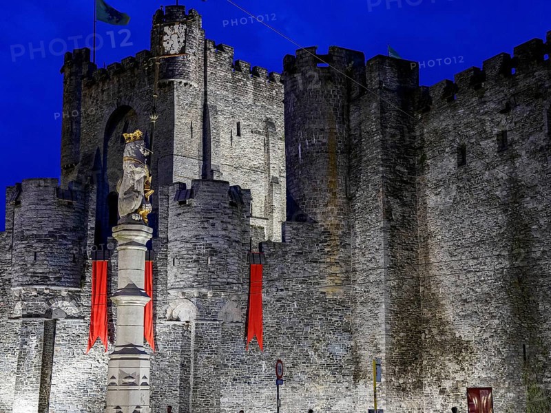 Medieval castle with massive stone walls, a statue and red flags illuminated at night, historic buildings with towers and bridges on a river at blue hour, gent, belgium