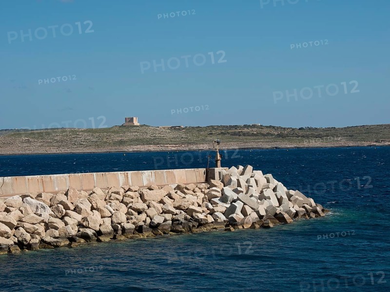 A breakwater of large stones in the sea with a rocky coastline in the background, the island of Gozo with historic houses, colourful balconies, magnificent churches and boats in the harbour, Gozo, Malta, Europe