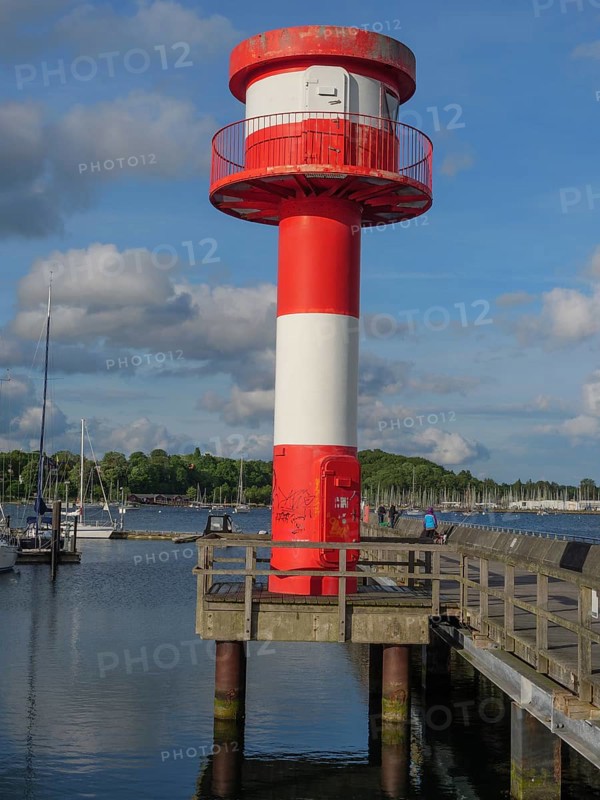 Close-up of a red and white lighthouse on a rough wooden jetty with blue sky and clouds, harbour with sailing boats and a small red and white lighthouse, Eckernfoerde, Germany, Europe