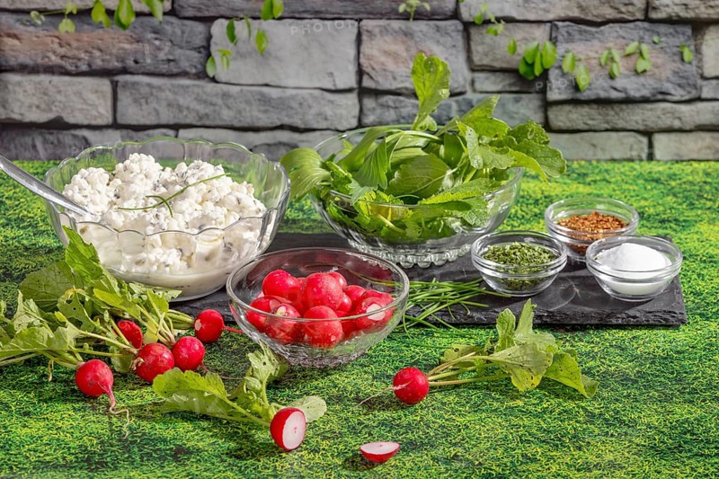 Radishes, cottage cheese, herbs and spices in glass bowls on grass in front of a stone wall