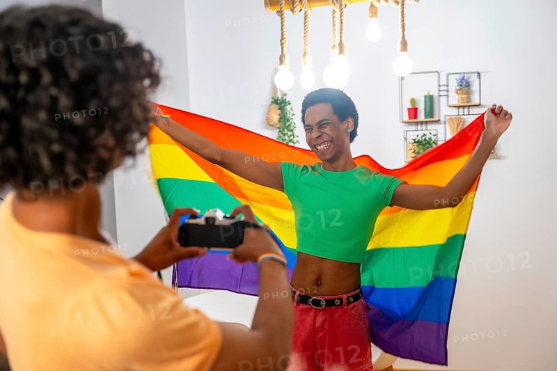 Gay friends taking pictures with camera while waving LGBT flag at home