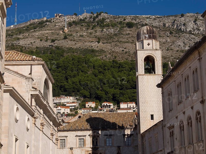 Church bell tower and historic buildings in front of a wooded mountain and blue sky, typical for summer days, the old town of Dubrovnik with historic houses, churches, red roofs and fortress walls, dubrovnik, coratia