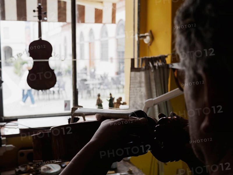 Silhouetted against a window, a luthier works on a stringed instrument in a workshop, with tools spread on the bench