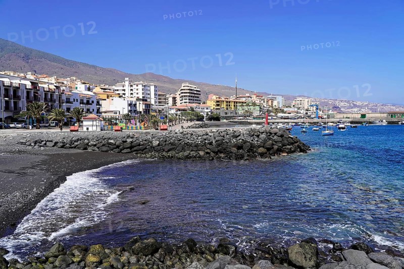 Black volcanic beach in Candelaria, Tenerife, Canary Islands, Spain, Europe, town on the coast with buildings, rocks and calm water under a blue sky, Europe