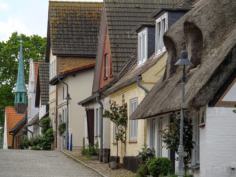Traditional houses with thatched roofs and a pointed tower at the end of the street, along a quiet, cobbled street in a European village, street with historic thatched houses in different colours, Maasholm, Germany, Europe