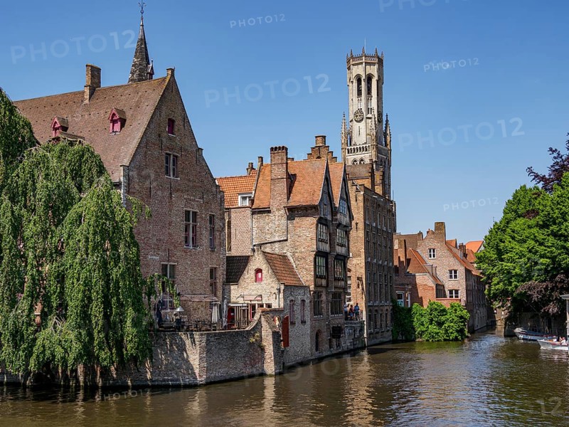 Historic buildings and a tall bell tower along a river on a sunny day, historic city by the river with ornate gables, old facades and historic towers, Bruges, Belgium, Europe