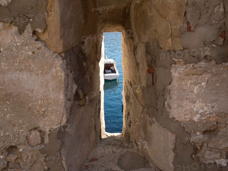 View through a narrow gap in an old stone wall to a boat in the sea, the old town of Dubrovnik with historic houses, churches, red roofs and fortress walls, dubrovnik, coratia