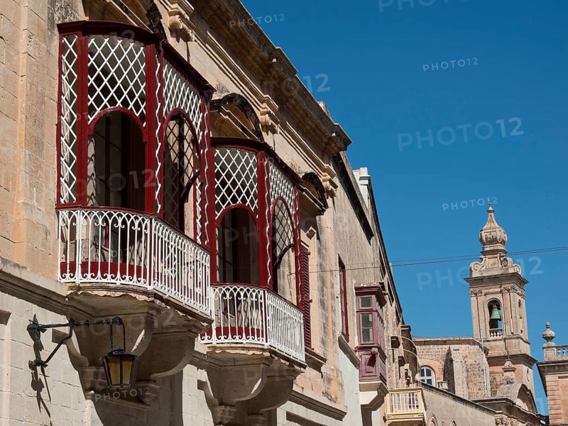 A view of an old town street with several balconies and a church tower in the background, decorative railings and Mediterranean architecture, old houses with balconies and towers, Gozo, Malta, Europe
