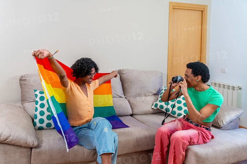 Gay man posing for his couple taking photos raising LGBT rainbow flag at home
