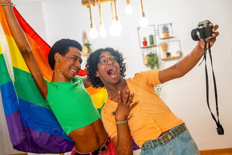 Gay latin young couple taking a selfie with a camera while raising rainbow lgbt flag at home