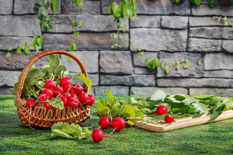 A basket of fresh radishes and a chopping board with herbs on a grassy area in front of a stone wall