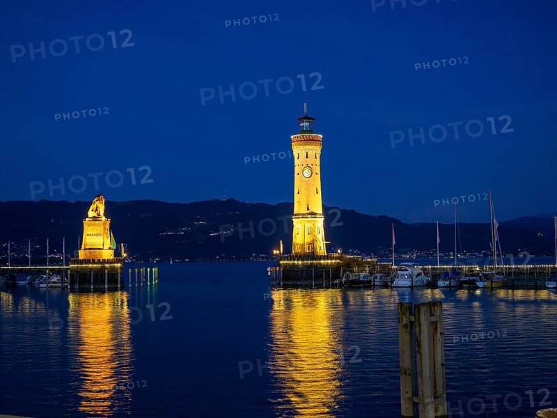 Illuminated lighthouse and lion statue at night, whose lights are reflected in the water of the harbour, towers at a harbour on the lake with sailing boats, Lindau, Germany, Europe